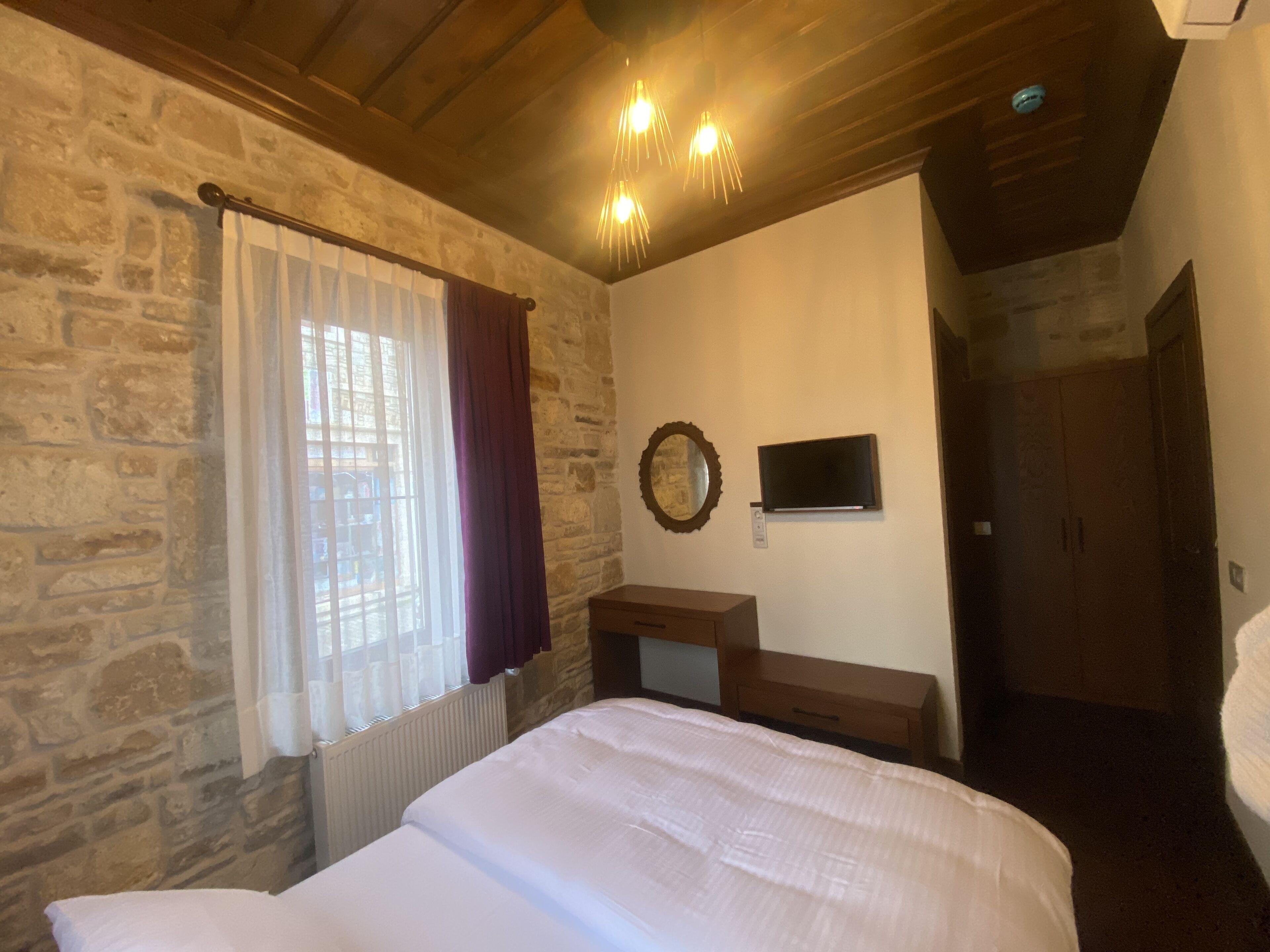 A bedroom with a stone wall, a bed, a dresser, a TV mounted on the wall, and a door leading to another room.