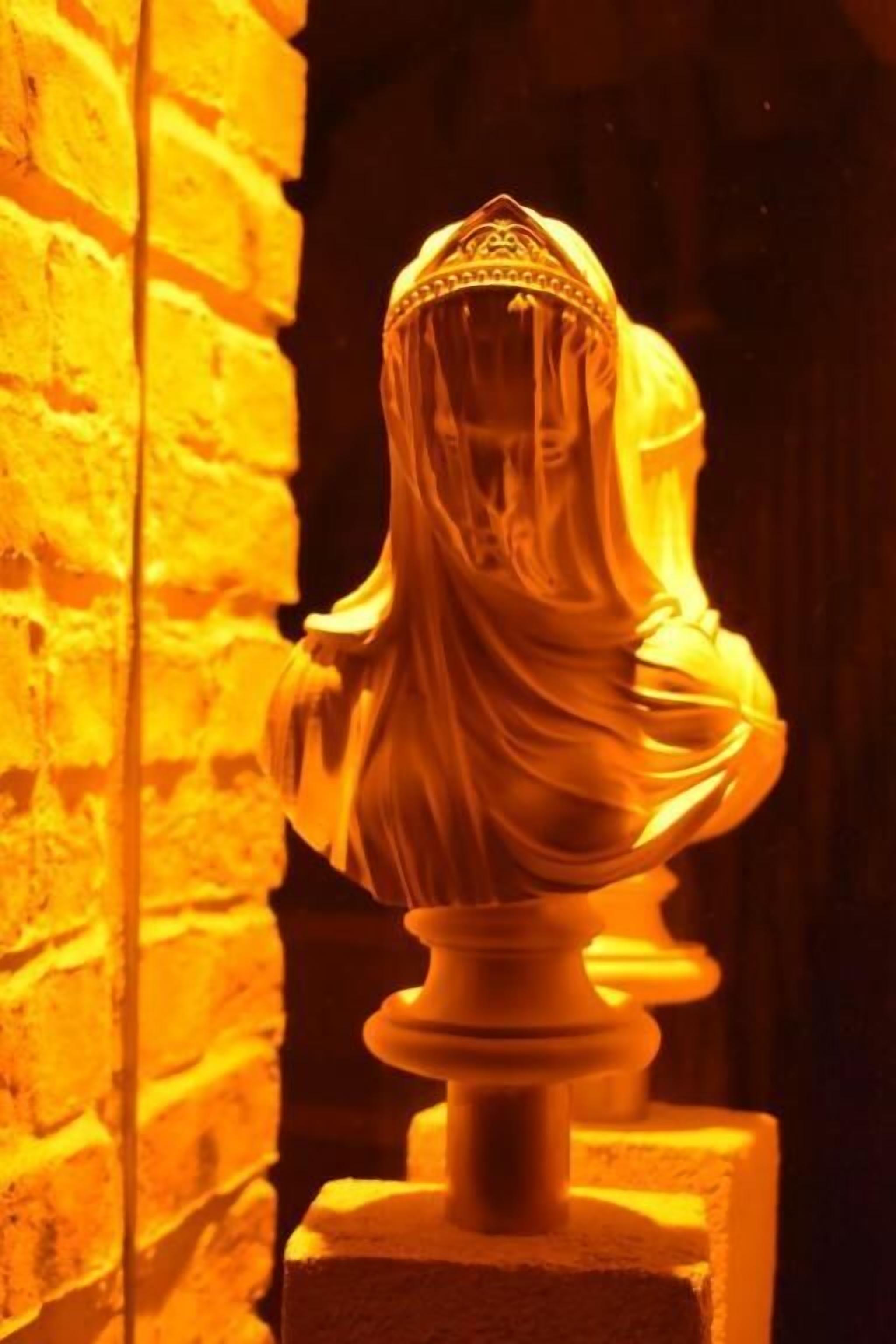 A bust sculpture with a hooded garment, set against a brick wall background.