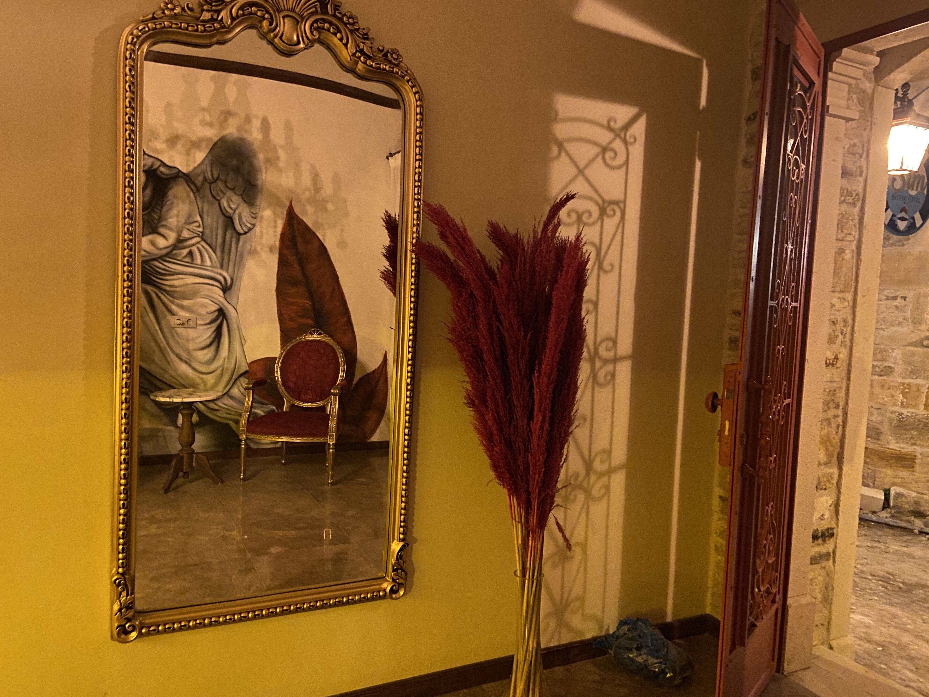 A mirror with a classical design, a red chair, and a decorative vase with red feathers.