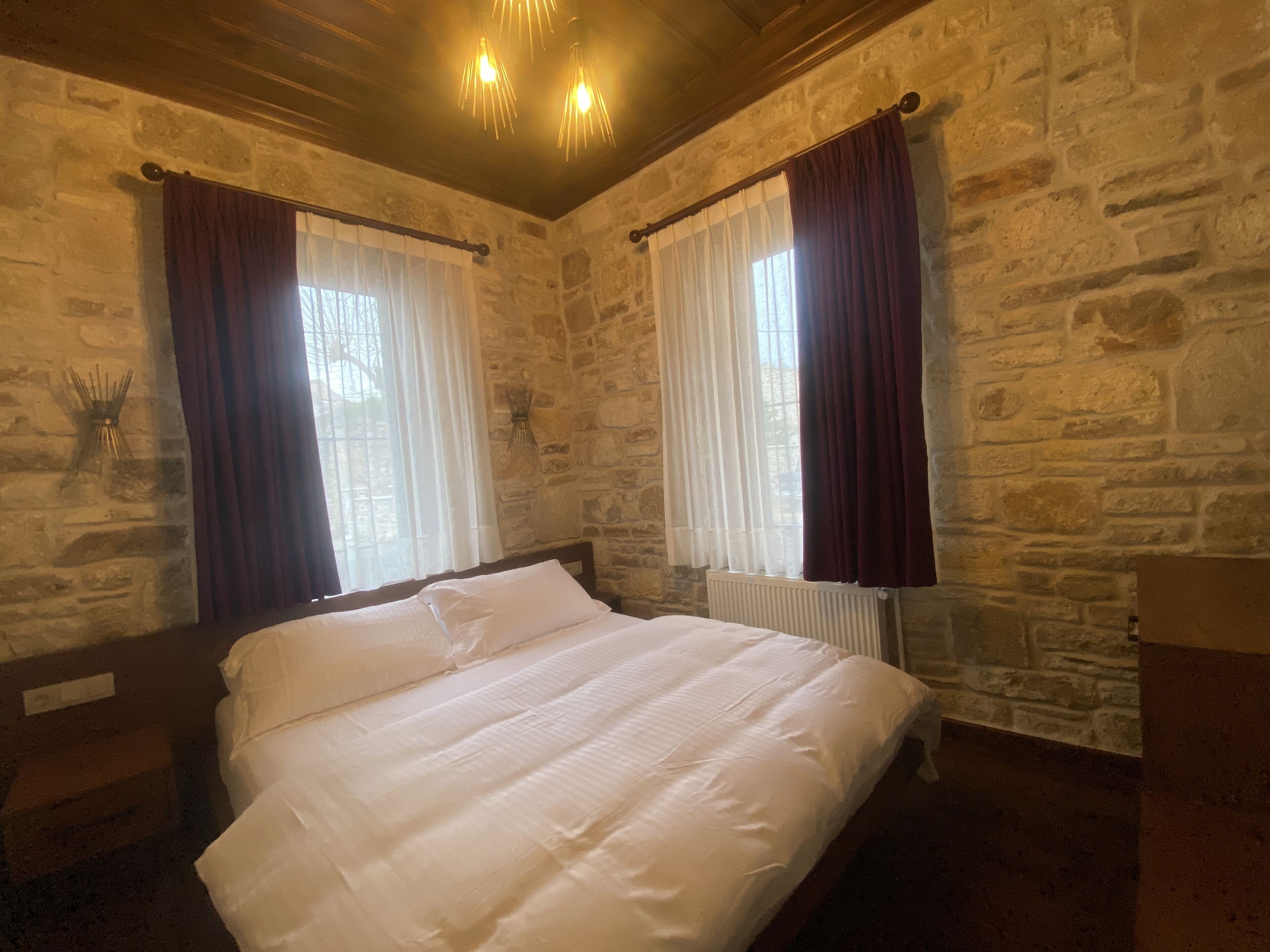 A room with a bed, stone walls, and two windows with curtains.