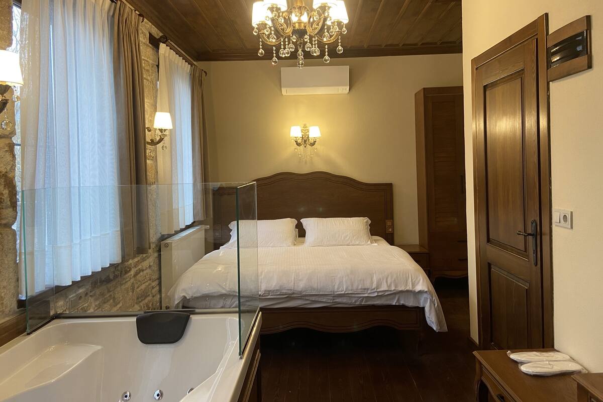 Quarto de hotel com uma cama grande, uma banheira de hidromassagem envidraçada, teto de madeira e um candelabro.