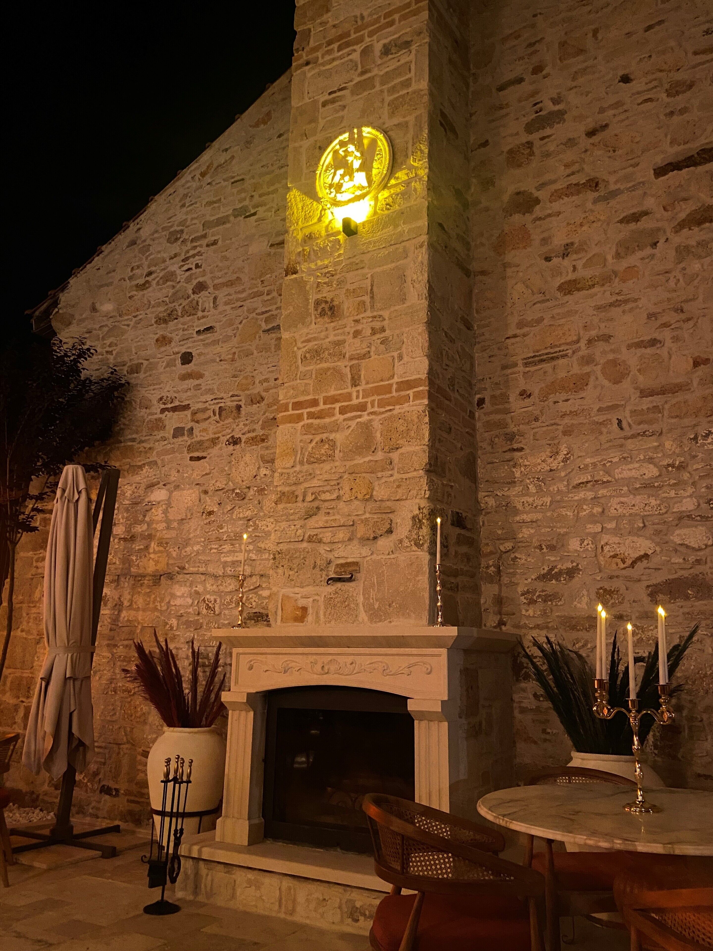 Un bâtiment en pierre avec un emblème illuminé au-dessus d’une cheminée, des places assises  à l’extérieur et une plante en pot.
