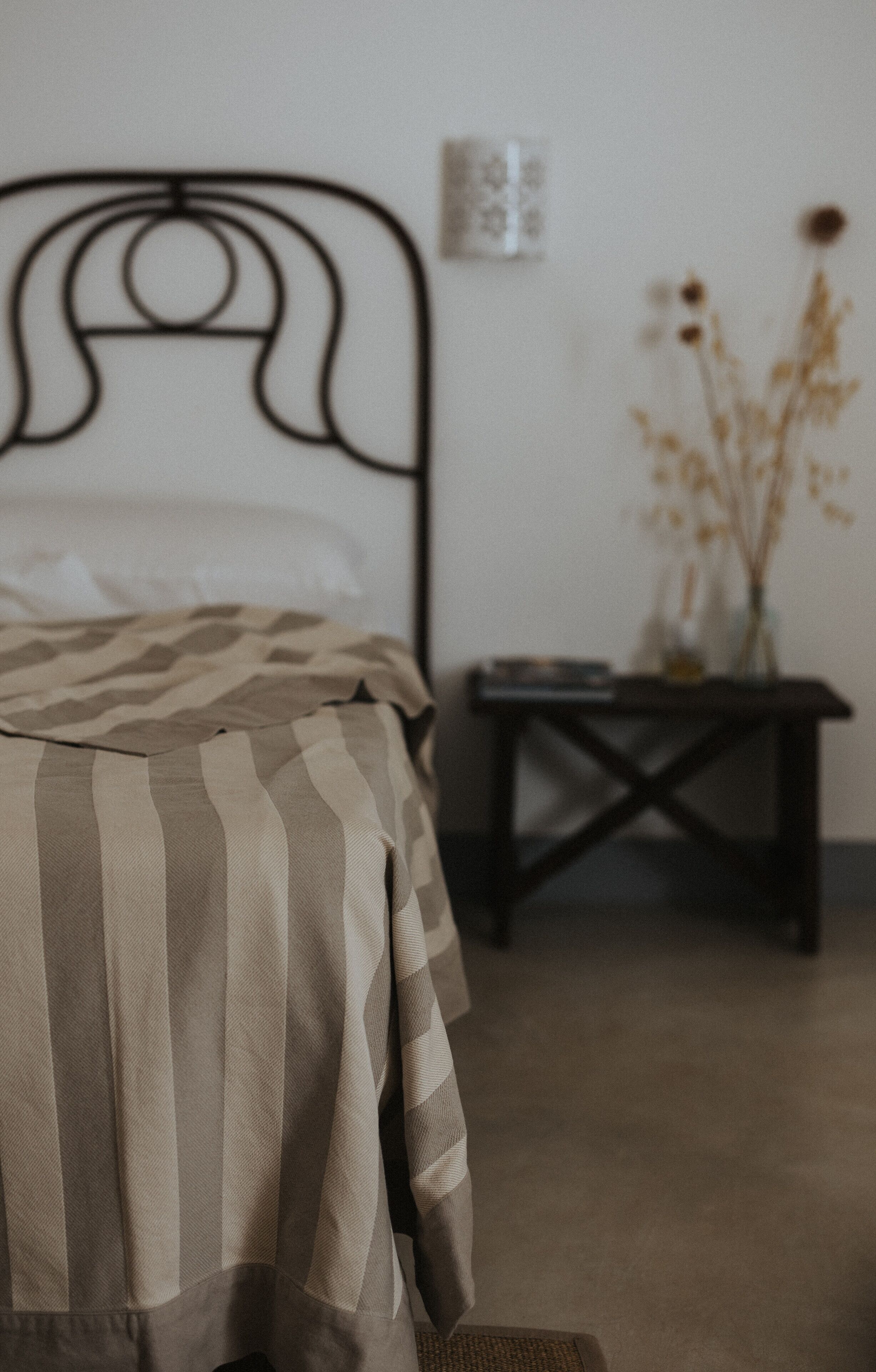 A bed with striped bedding, a dark metal headboard, a small bedside table with a vase of dried flowers, and a wall-mounted light fixture.