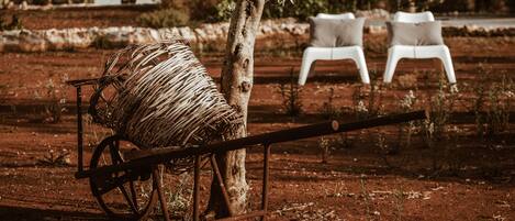 A rustic outdoor setting with a wooden cart loaded with a large bundle of straw, a tree, and two white plastic chairs in the background.