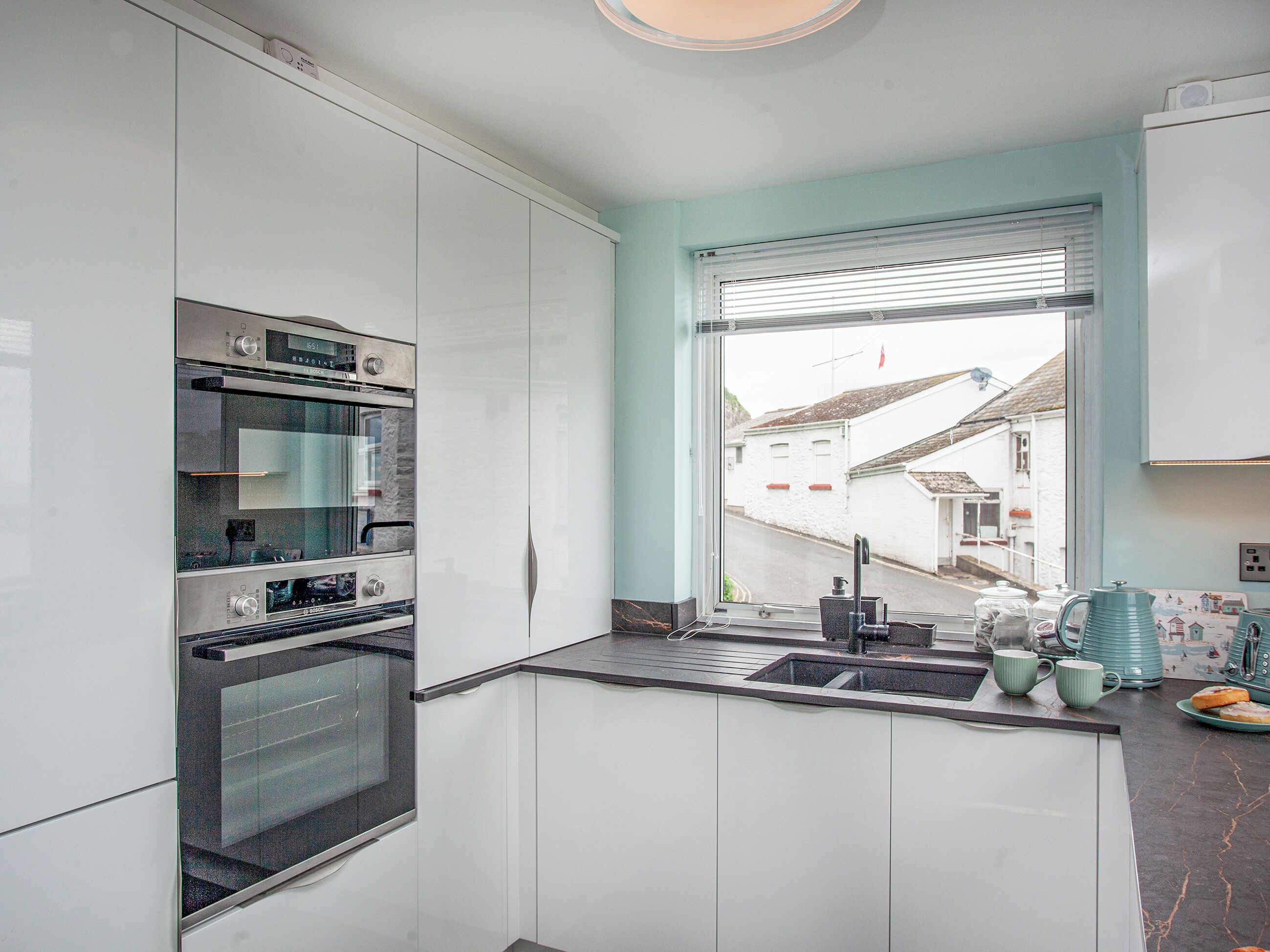 A modern kitchen with white cabinets, a built-in oven, a sink, and a window overlooking a street.