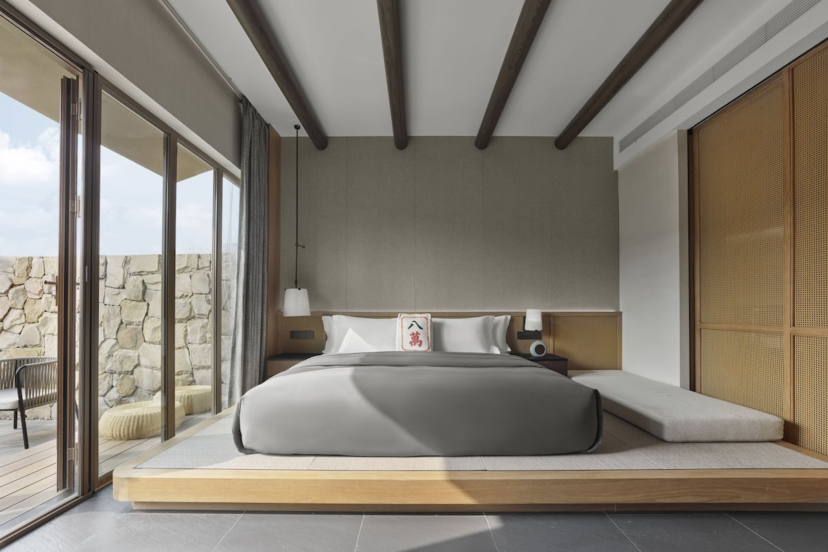 A modern bedroom with a large bed, a stone wall, and a view of the outdoors.