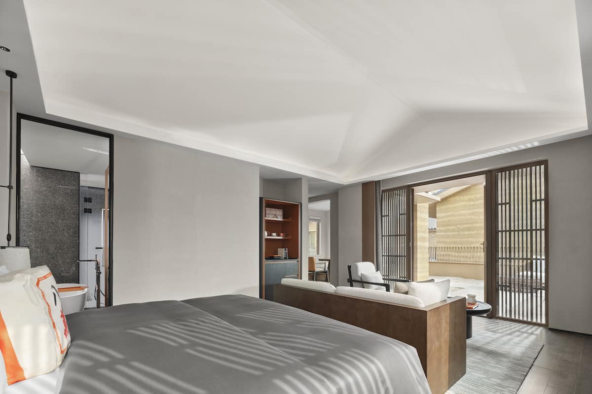A modern hotel room with a large bed, a sitting area with a sofa and armchairs, a small desk, and a view of the outdoor balcony.