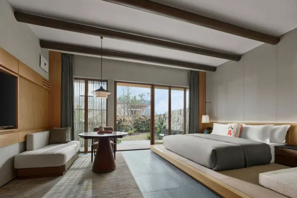 A modern hotel room with a large bed, a sofa, a dining table, and a view of the outdoors.