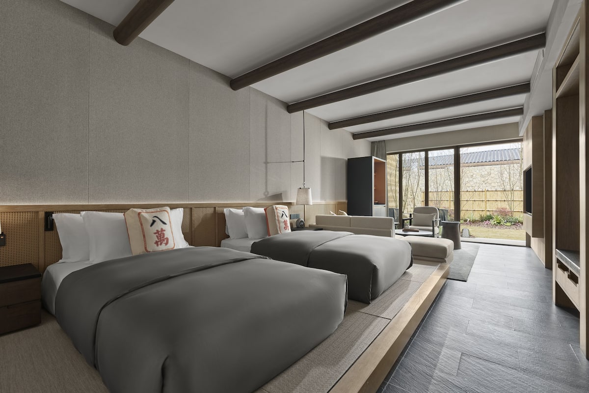 A modern hotel room with two beds, a sofa, a TV, and a large window offering a view of the outdoors.