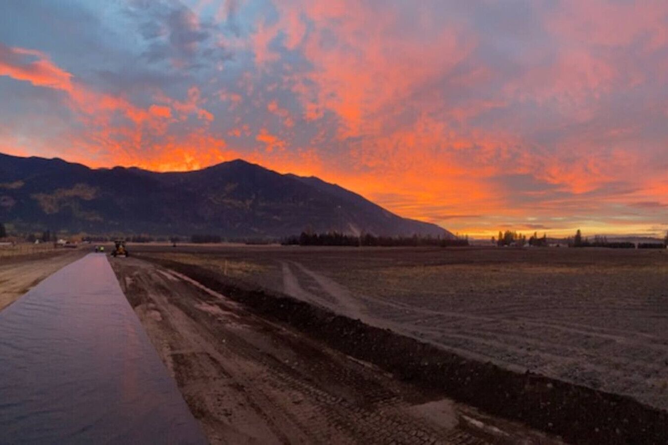 Een landweg met een verhard pad aan de linkerkant en een onverharde weg aan de rechterkant, die leidt naar een bergketen onder een kleurrijke zonsondergang.