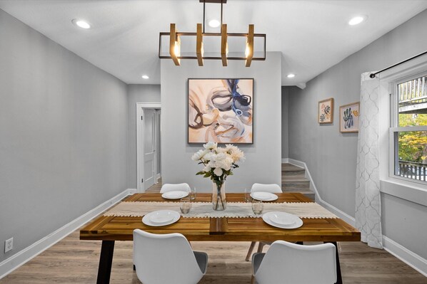 A dining area with a wooden table, white chairs, a vase with flowers, and a modern chandelier. - Newly remodeled 4BRs next to OSU&Expo, 2 FREE Parking. (Columbus)