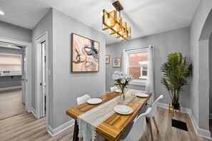 A dining area with a wooden table, white chairs, a white lace table runner, a vase with flowers, and a modern pendant light. - Newly remodeled 4BRs next to OSU&Expo, 2 FREE Parking. (Columbus)