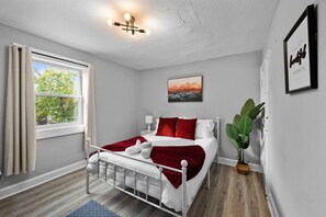 A bedroom with a bed, a window with curtains, a bedside table, a wall-mounted picture, and a potted plant. - Newly remodeled 4BRs next to OSU&Expo, 2 FREE Parking. (Columbus)