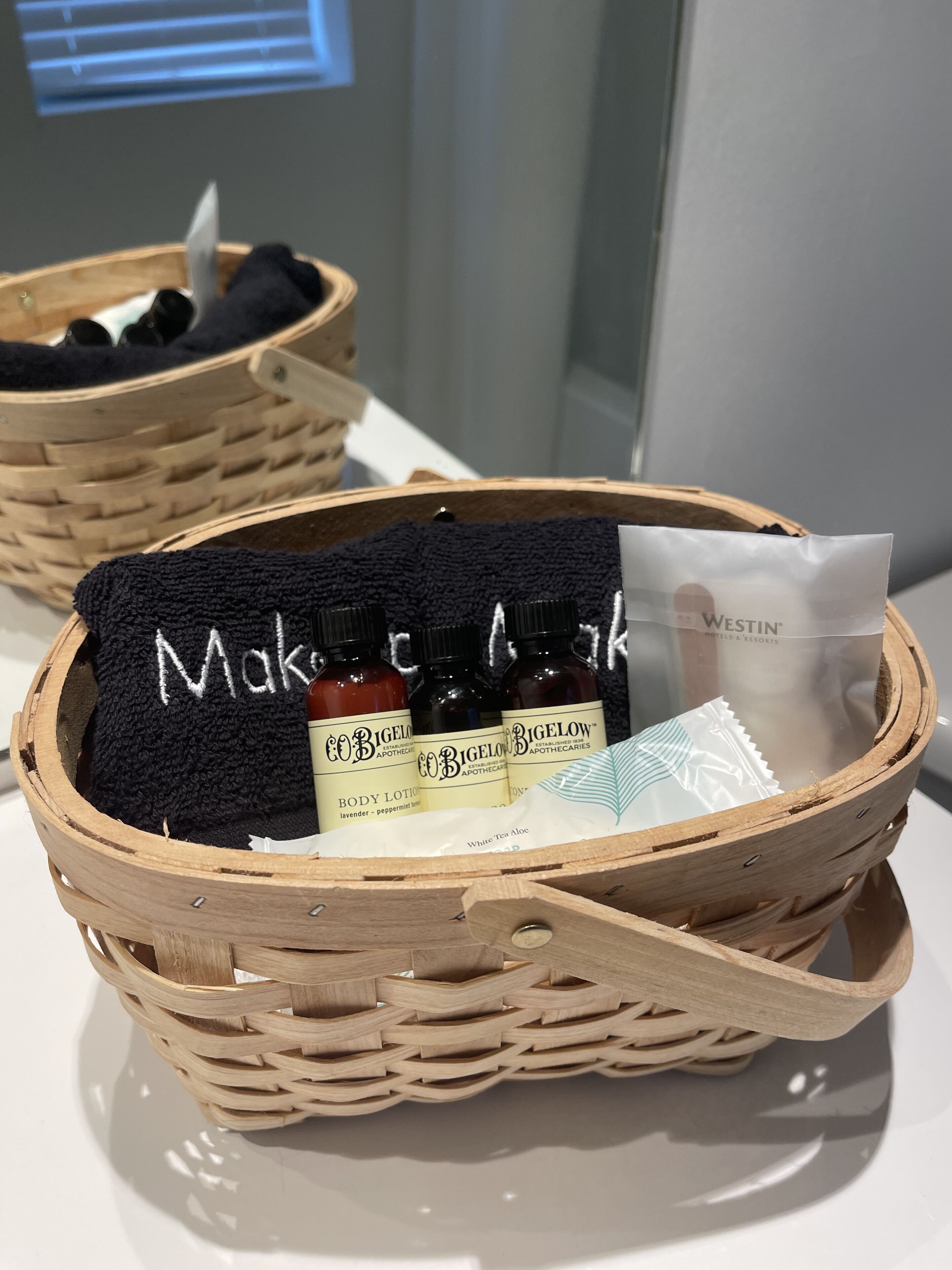 Two wicker baskets containing toiletries and a black towel with "Make" embroidered on it.