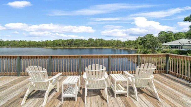 Um deck de madeira com quatro cadeiras Adirondack brancas voltado para um lago, cercado por um corrimão de madeira.