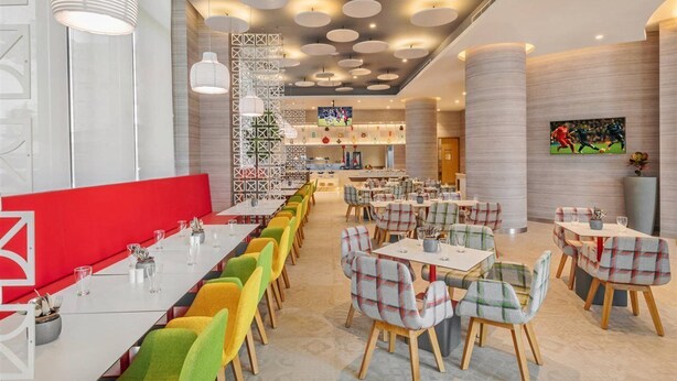A modern restaurant with multiple dining areas, including a long communal table and smaller tables with plaid chairs. - Ibis Kolkata Rajarhat (Barasat)