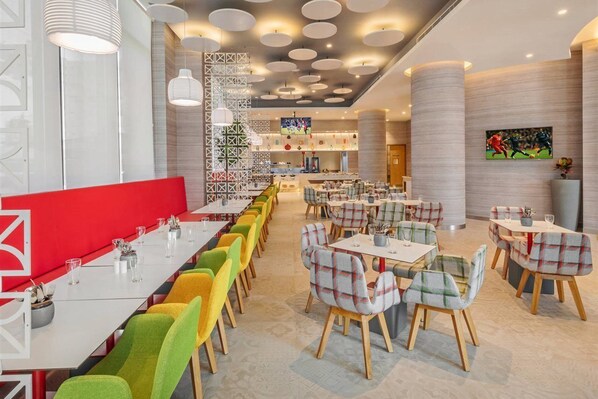 A modern restaurant with multiple dining areas, including a long communal table and smaller tables with plaid chairs. - Ibis Kolkata Rajarhat (Barasat)