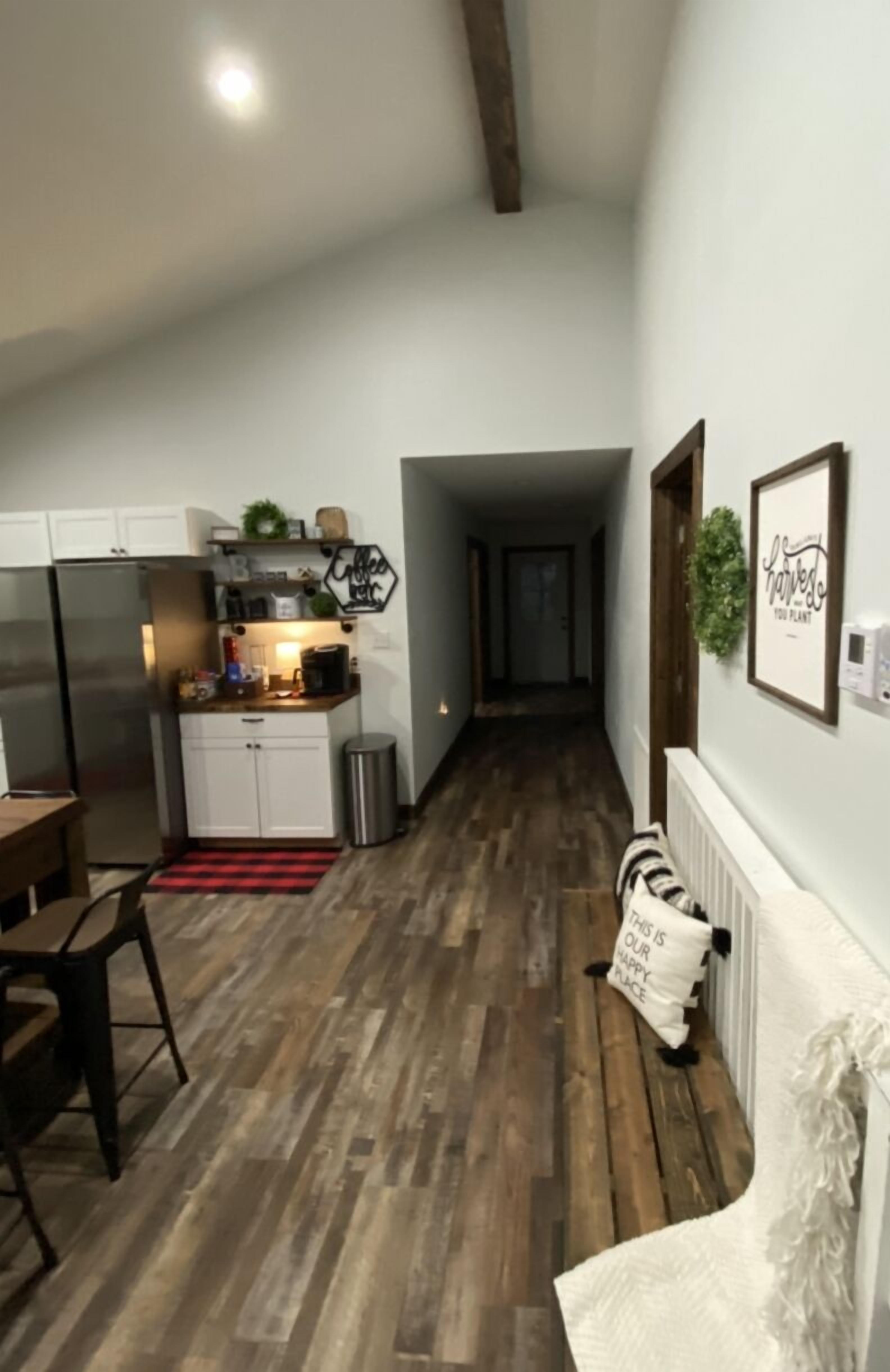 A narrow hallway with a kitchen area on one side, featuring white cabinets, a stainless steel refrigerator, and a countertop with various items.