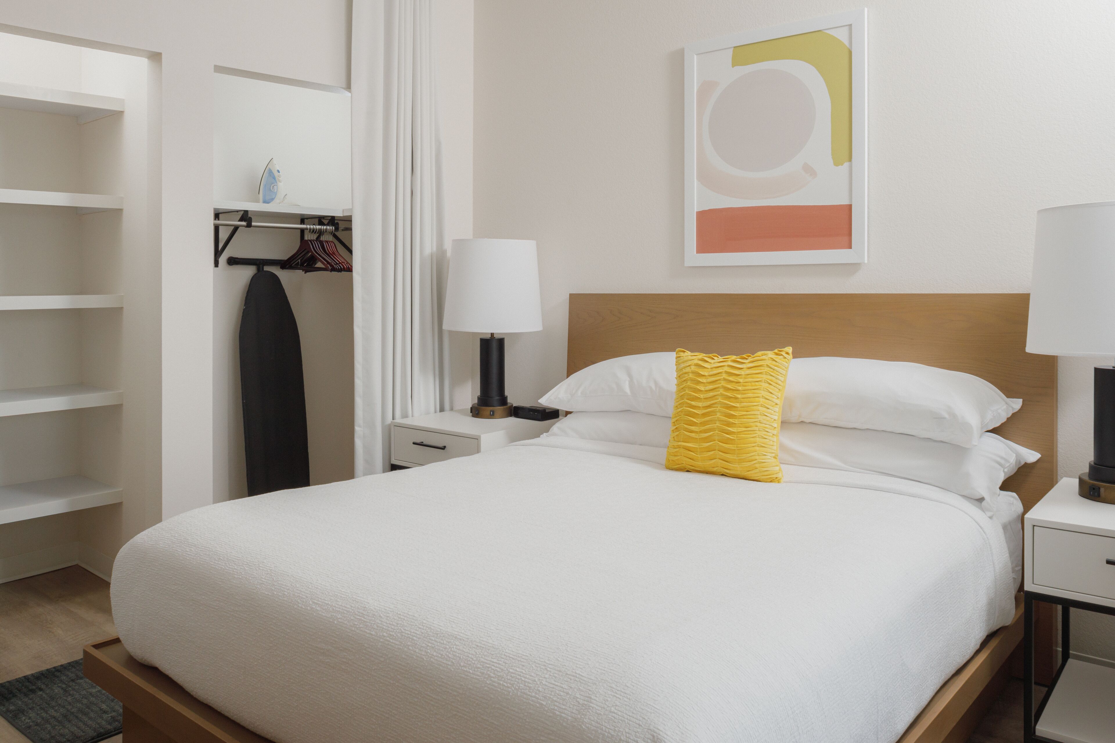 Una cama bien hecha con edredón blanco y almohada amarilla decorativa, una mesita de noche con lámpara, un perchero y una obra de arte abstracto colgada en la pared.