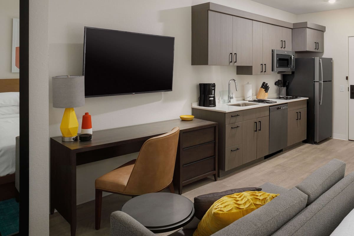 Una habitación de hotel moderna con una televisión de pantalla plana, un escritorio con una silla, un sofá y una zona de cocina con microondas y refrigerador.