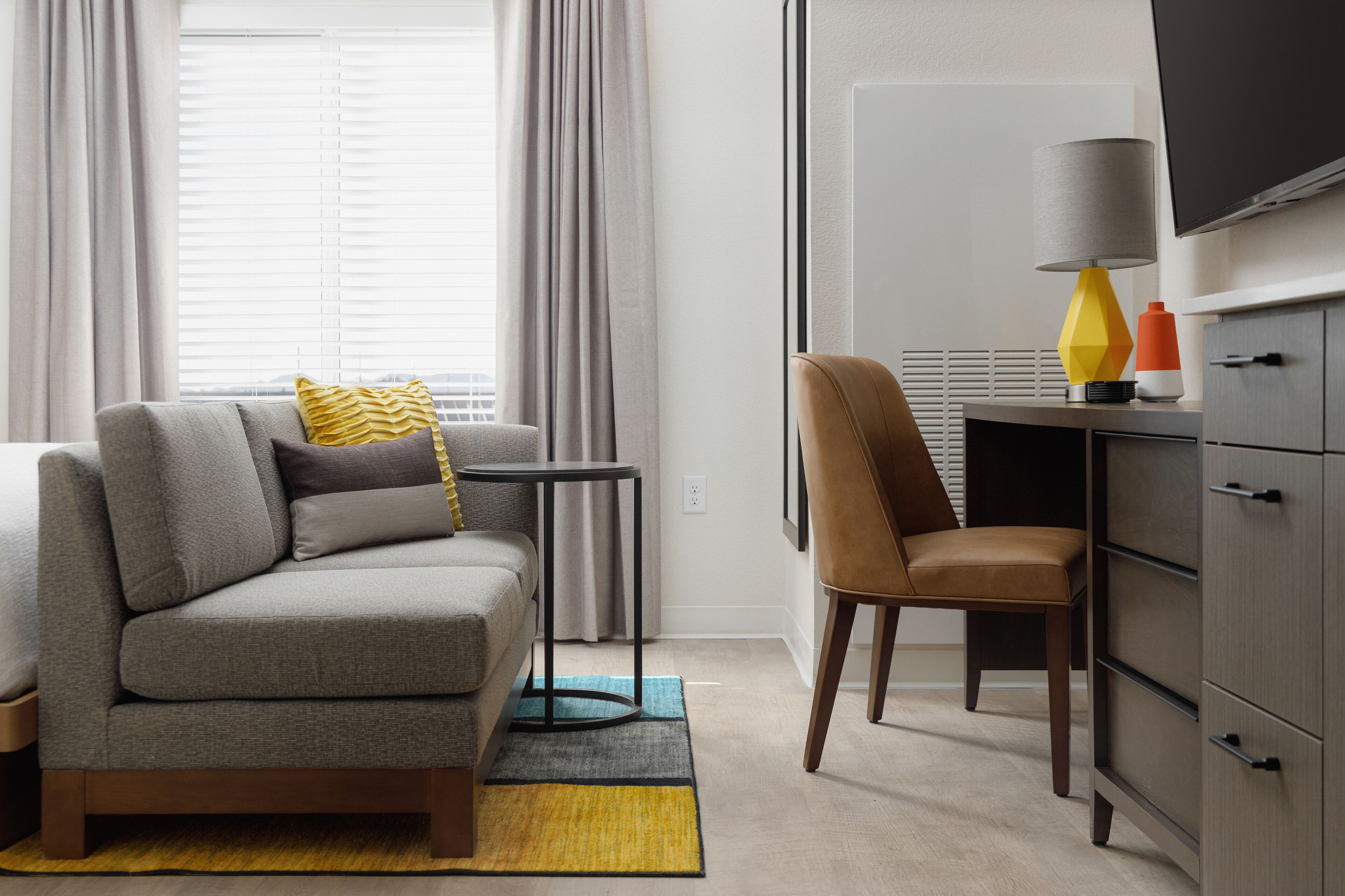 Quarto de hotel moderno com um sofá cinzento, uma cadeira marrom, uma mesa pequena, uma luminária e uma TV sobre um móvel.