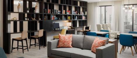 A modern living room with a grey sofa, orange patterned pillows, a bookshelf, and a dining area with blue chairs.