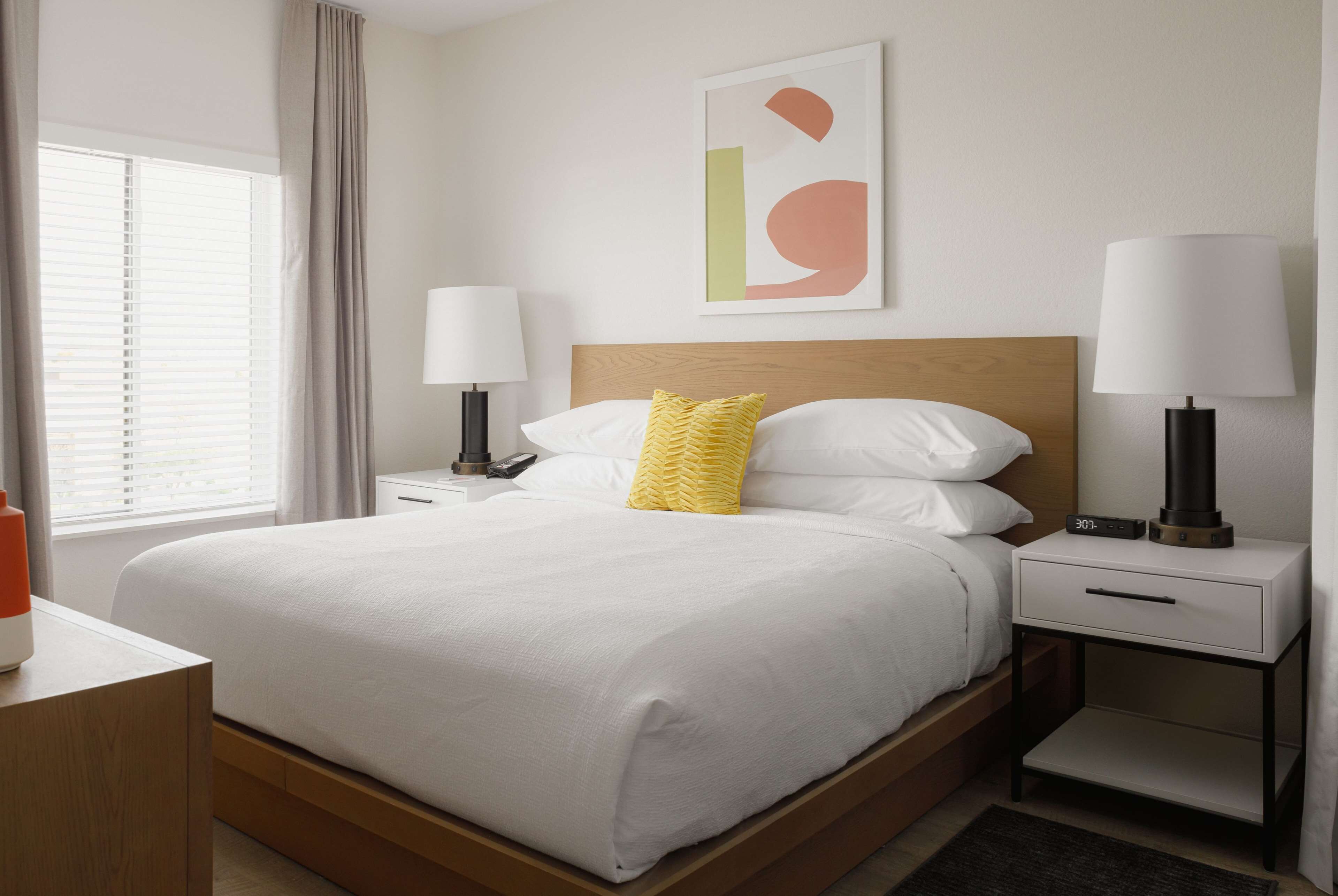 Een keurig opgemaakt hotelkamer met een groot bed, nachtkastjes met lampen, een wekker en een raam met jaloezieën.