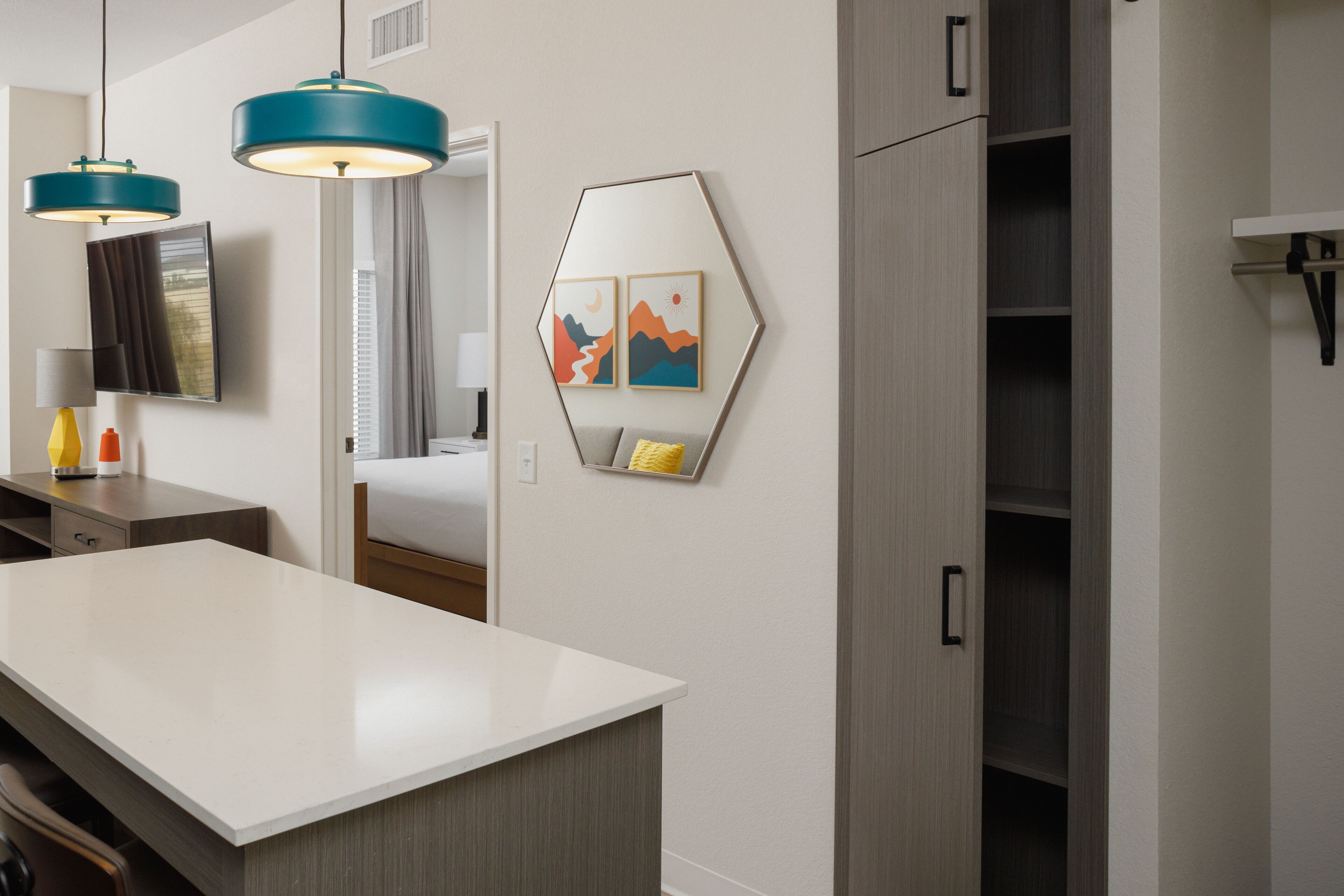 A modern hotel room with a large white desk, two teal pendant lights, a bed with a view, a wardrobe, and a wall-mounted TV.