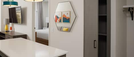 A modern hotel room with a large white desk, two teal pendant lights, a bed with a view, a wardrobe, and a wall-mounted TV.