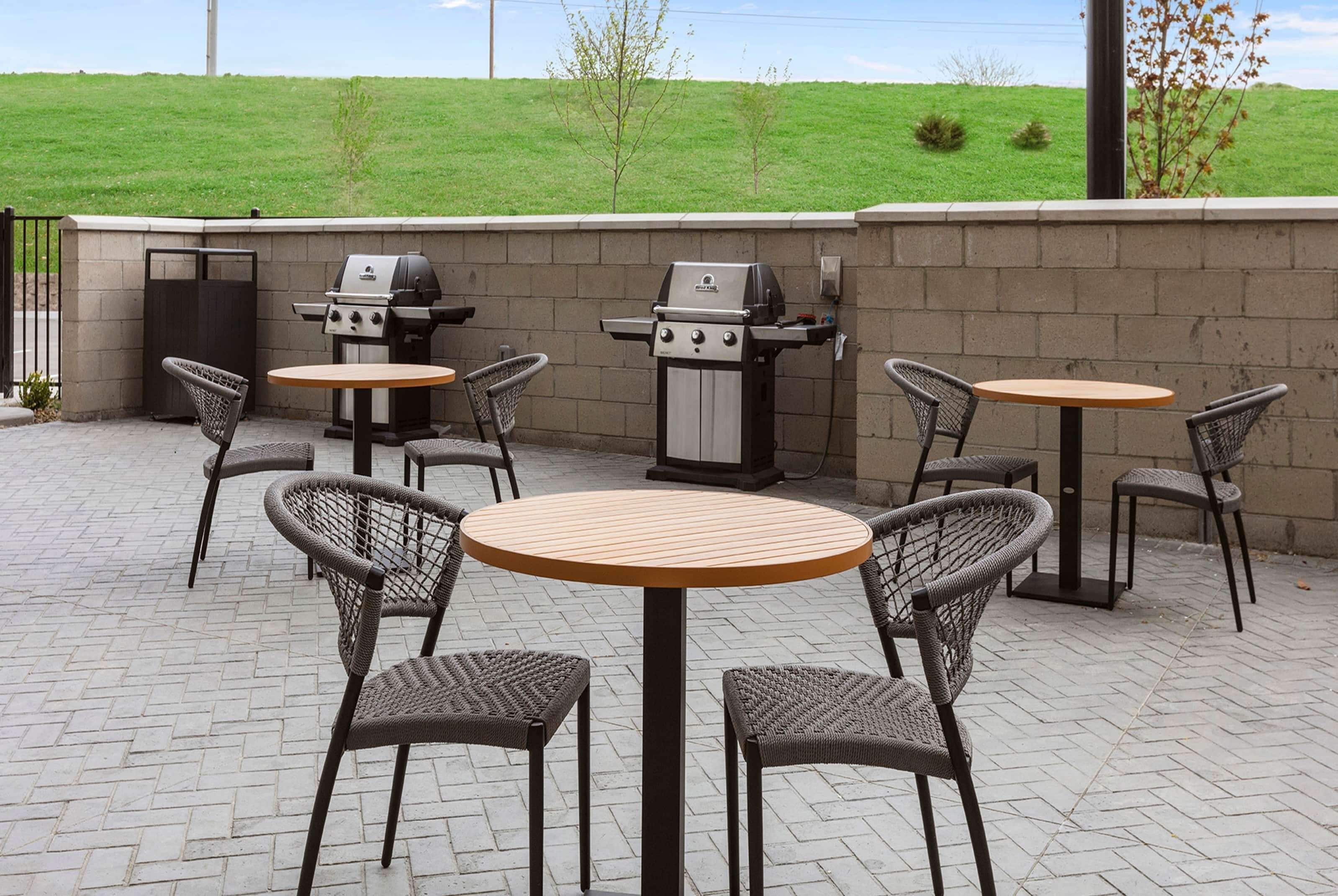 Une terrasse extérieure avec quatre tables rondes et des chaises en osier, un barbecue et un mur en briques.
