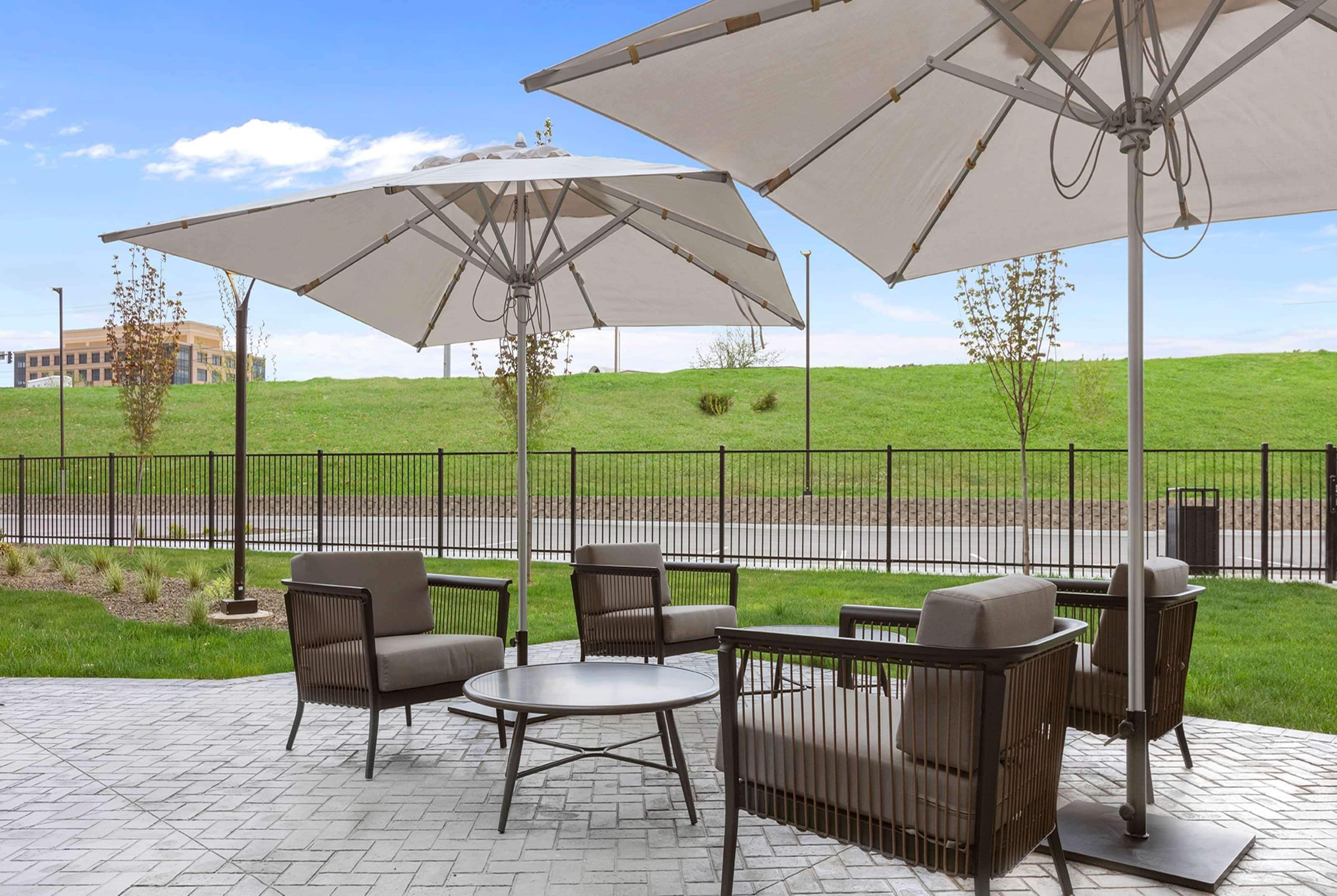 Eine Terrasse im Freien mit Sitzgelegenheiten und Sonnenschirmen, ein sanfter Hügel im Hintergrund und ein Zaun.