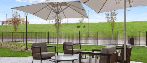 An outdoor patio with seating and umbrellas, a grassy hill in the background, and a fence.