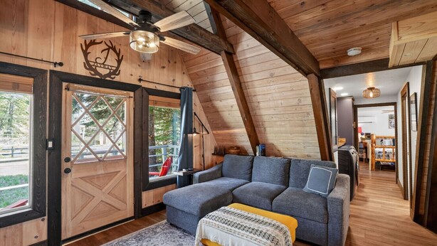 Ein gemütliches Loft-Zimmer mit Holzbalkendecke, großem Fenster, grauem Sofa und einer kleinen, gelben Bank.
