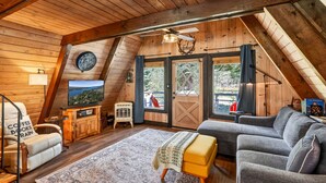 Ein gemütliches Dachzimmer mit Holzwänden, einem großen Fenster, einer Couch, einem Couchtisch und einem Fernseher.