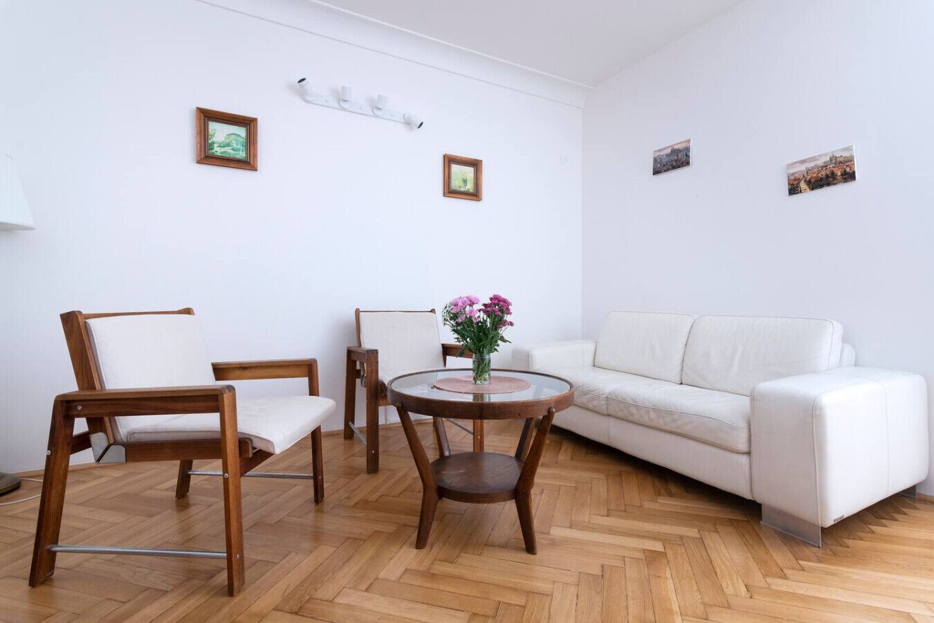 Een woonkamer met een witte bank, twee houten stoelen, een ronde tafel met een bloevas en twee ingelijste foto's aan de muur.