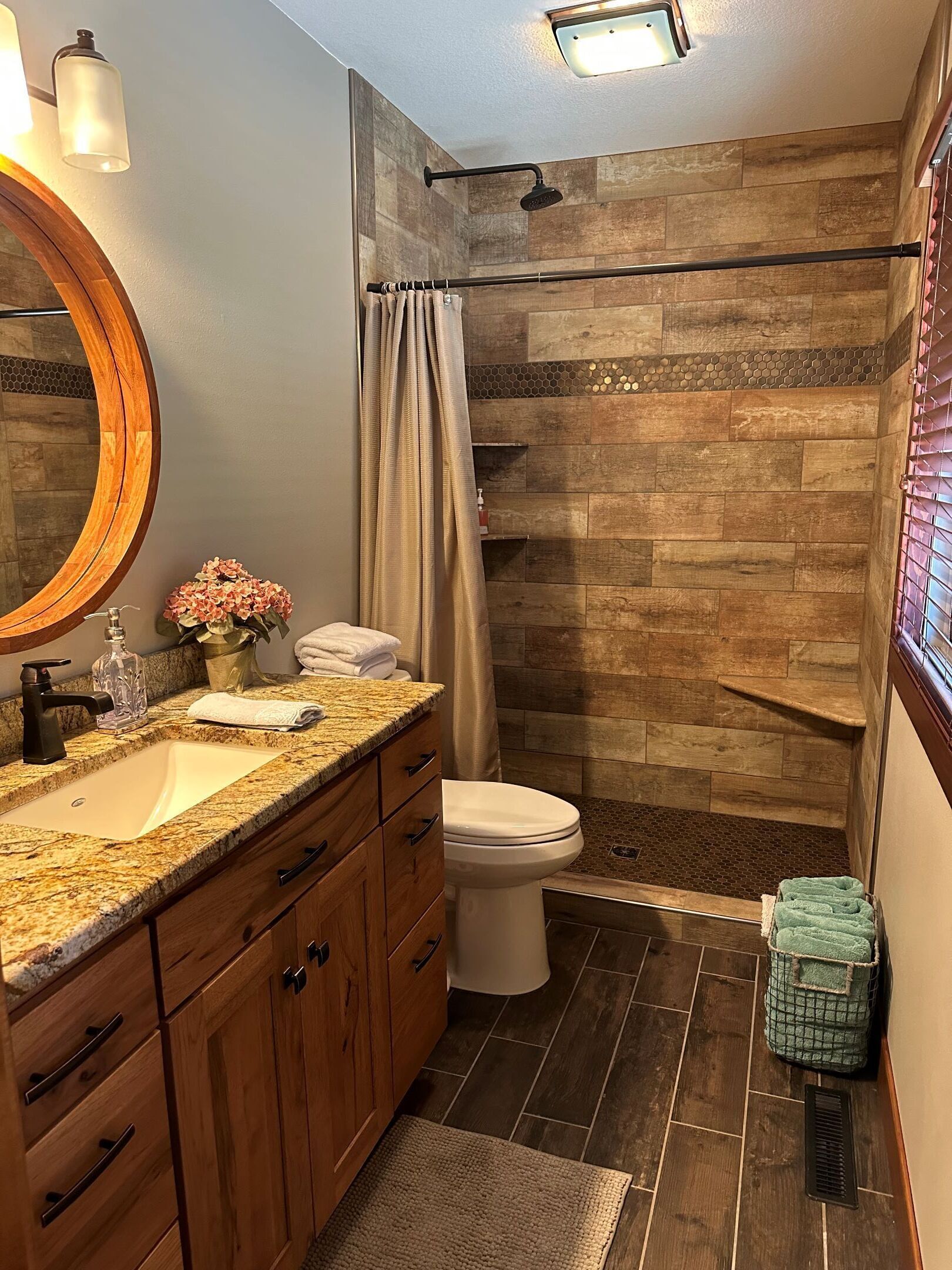 Une salle de bain avec un lavabo sur un plan de travail en granit, des meubles en bois, des toilettes blanches et une douche avec un mur en bois.