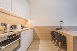 Una cocina moderna con horno empotrado, cafetera y hervidor. Hay una encimera de madera y una pequeña zona de comedor con dos sillas.