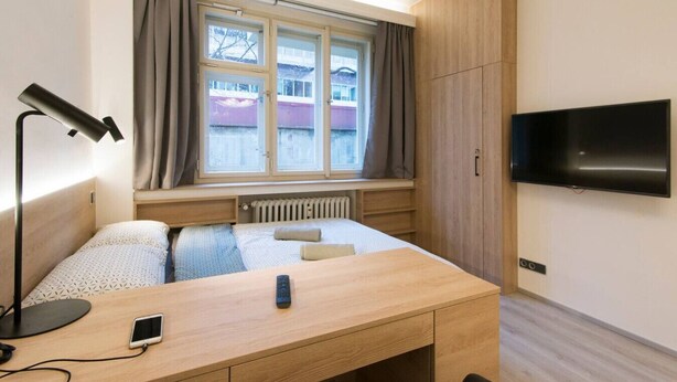 A hotel room with a bed, desk, lamp, TV, and window with curtains. - Quiet Studio by Anděl Metro - Walk to Old Town, Next to Nový Smíchov Mall (Prague)