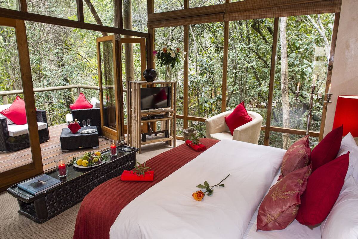 Un dormitorio con una cama grande, almohadas rojas, una mesita de noche con velas y vistas al bosque a través de amplias ventanas.