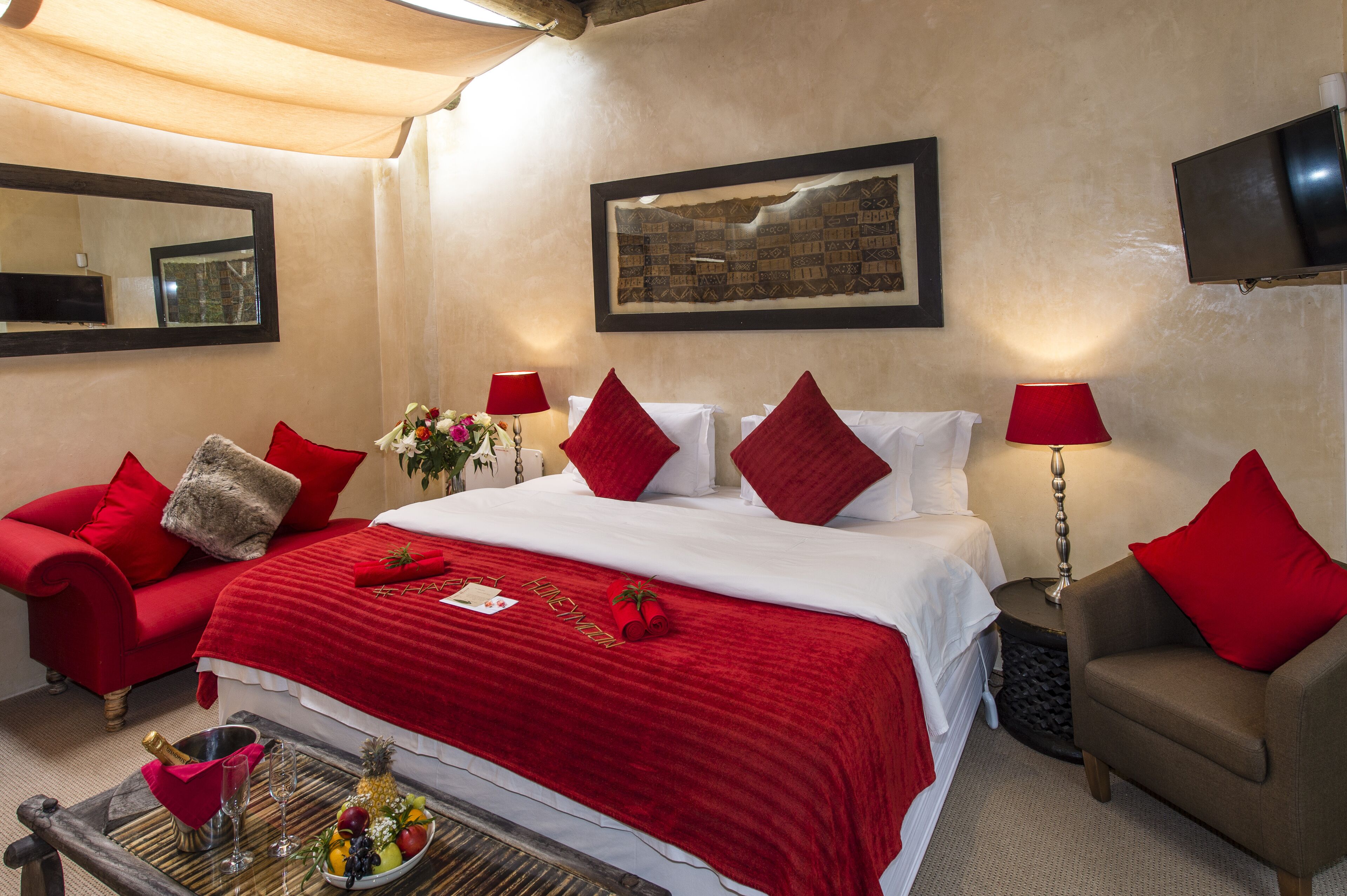 A hotel room with a bed, red and white bedding, a red armchair, a small table with a fruit bowl, a bottle of champagne, and a mirror on the wall.
