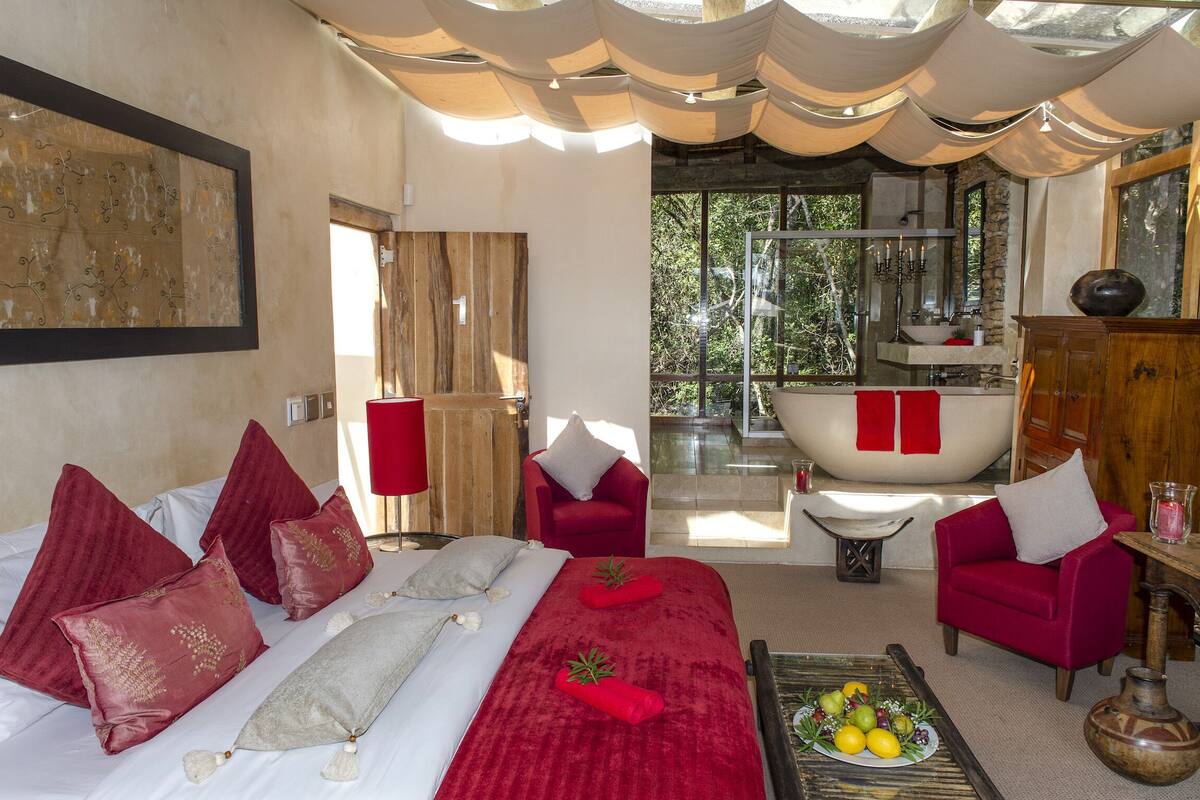 Un dormitorio con una cama grande, almohadas rojas y blancas, una silla roja, un armario de madera y vistas a un baño con bañera.