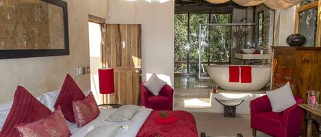 A bedroom with a large bed, red and white pillows, a red chair, a wooden cabinet, and a view of a bathroom with a bathtub.
