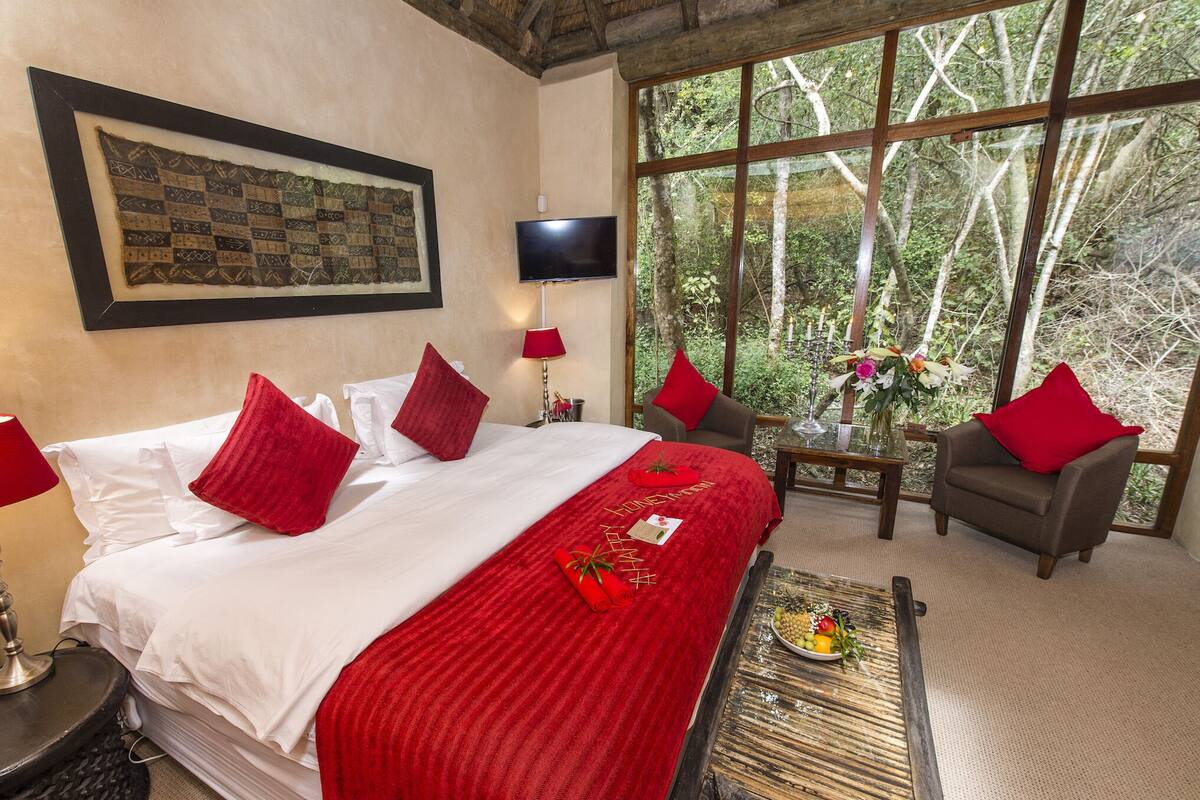Habitación de hotel con una cama grande, almohadas rojas, un televisor de pantalla plana, una mesita con fruta y vistas a árboles a través de ventanas amplias.