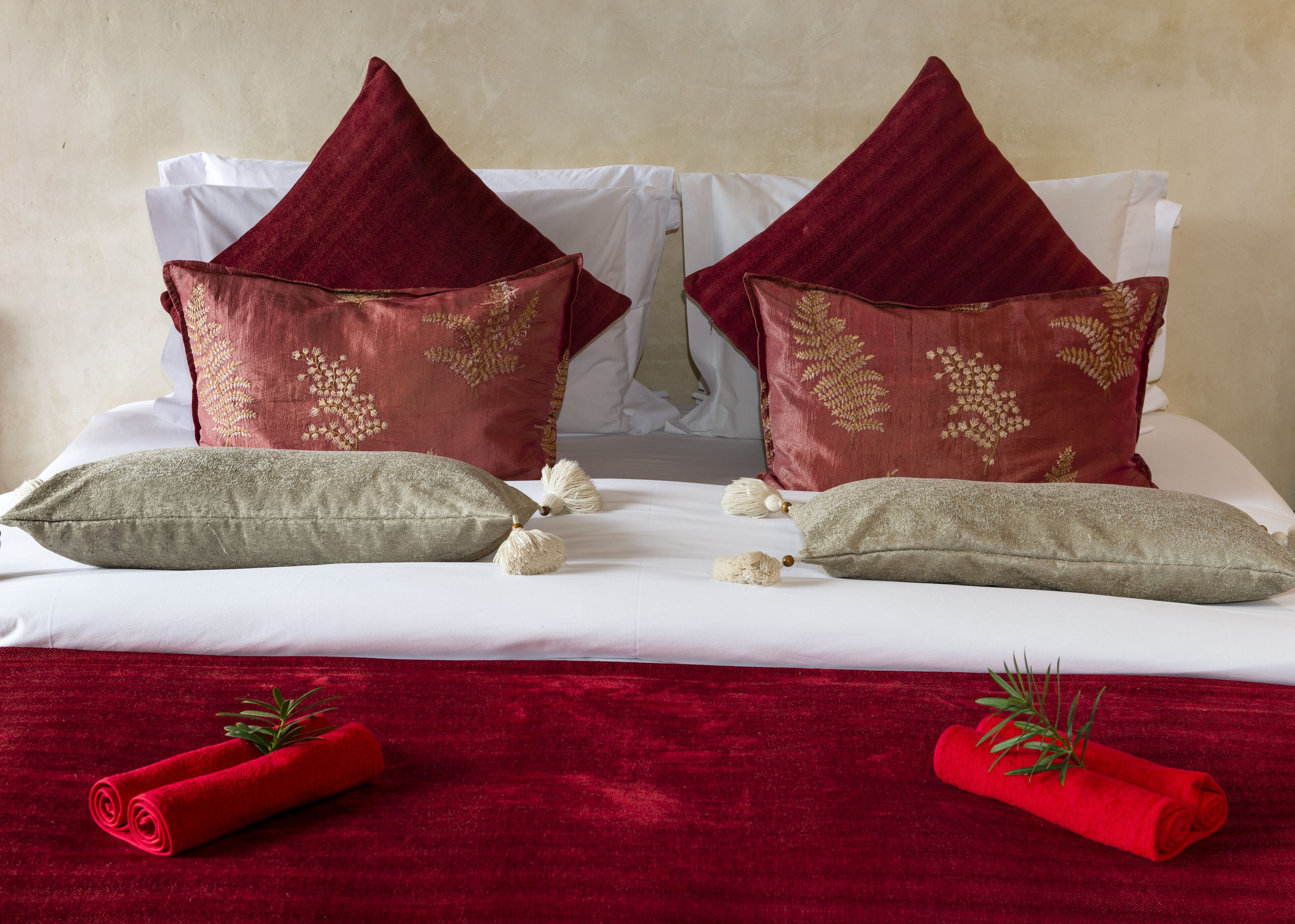 A bed with two red pillows with gold embroidery, two beige pillows, and a red towel with a sprig of rosemary.