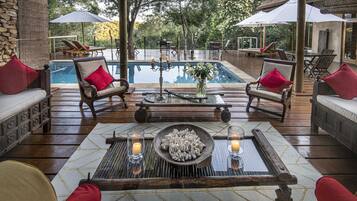 Un área junto a la piscina con mobiliario de madera, una mesa con velas y vistas a la piscina y árboles.