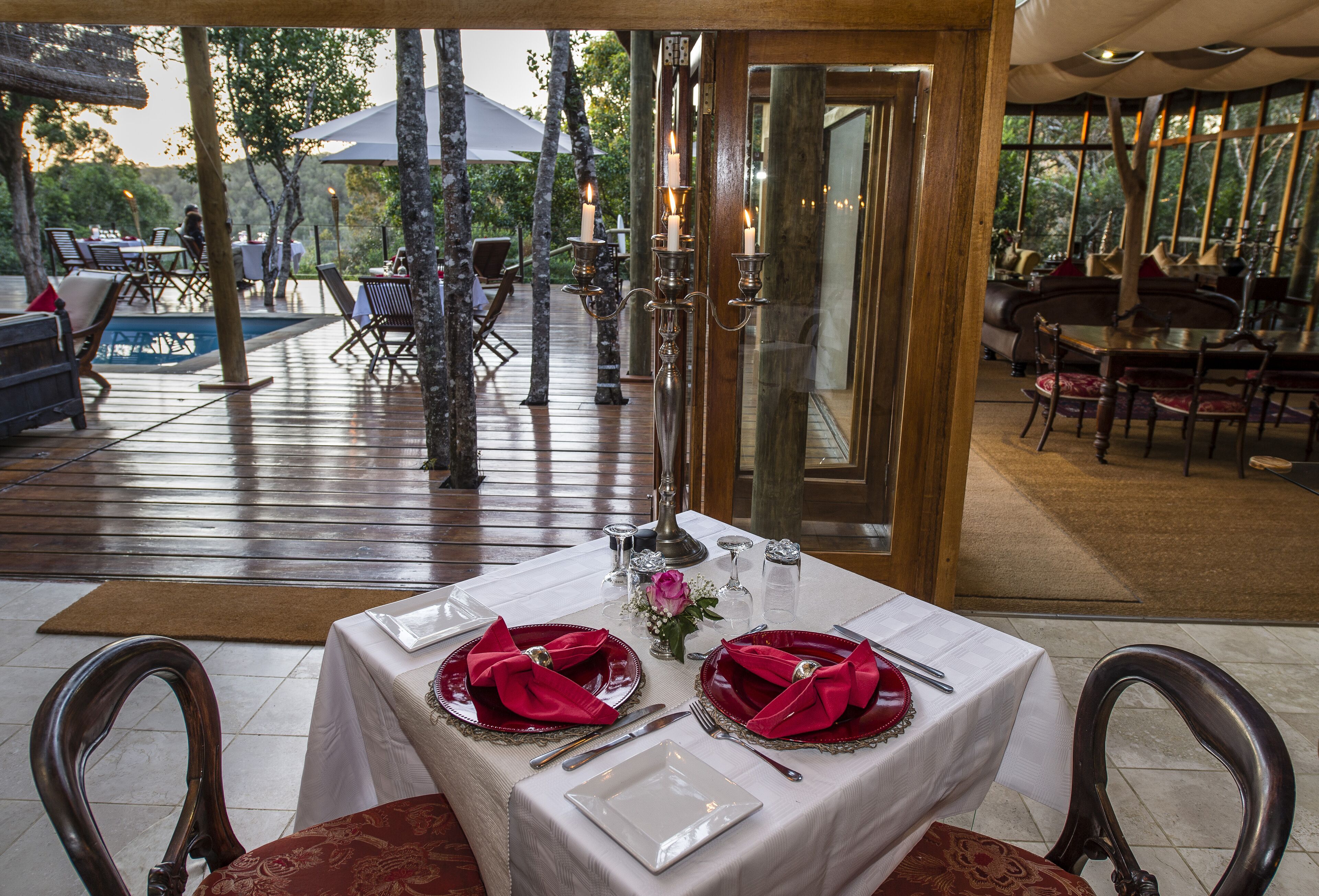 Zona pranzo all'aperto con tavolo apparecchiato per due, circondata da sedie di legno, una piscina e un pannello di vetro.