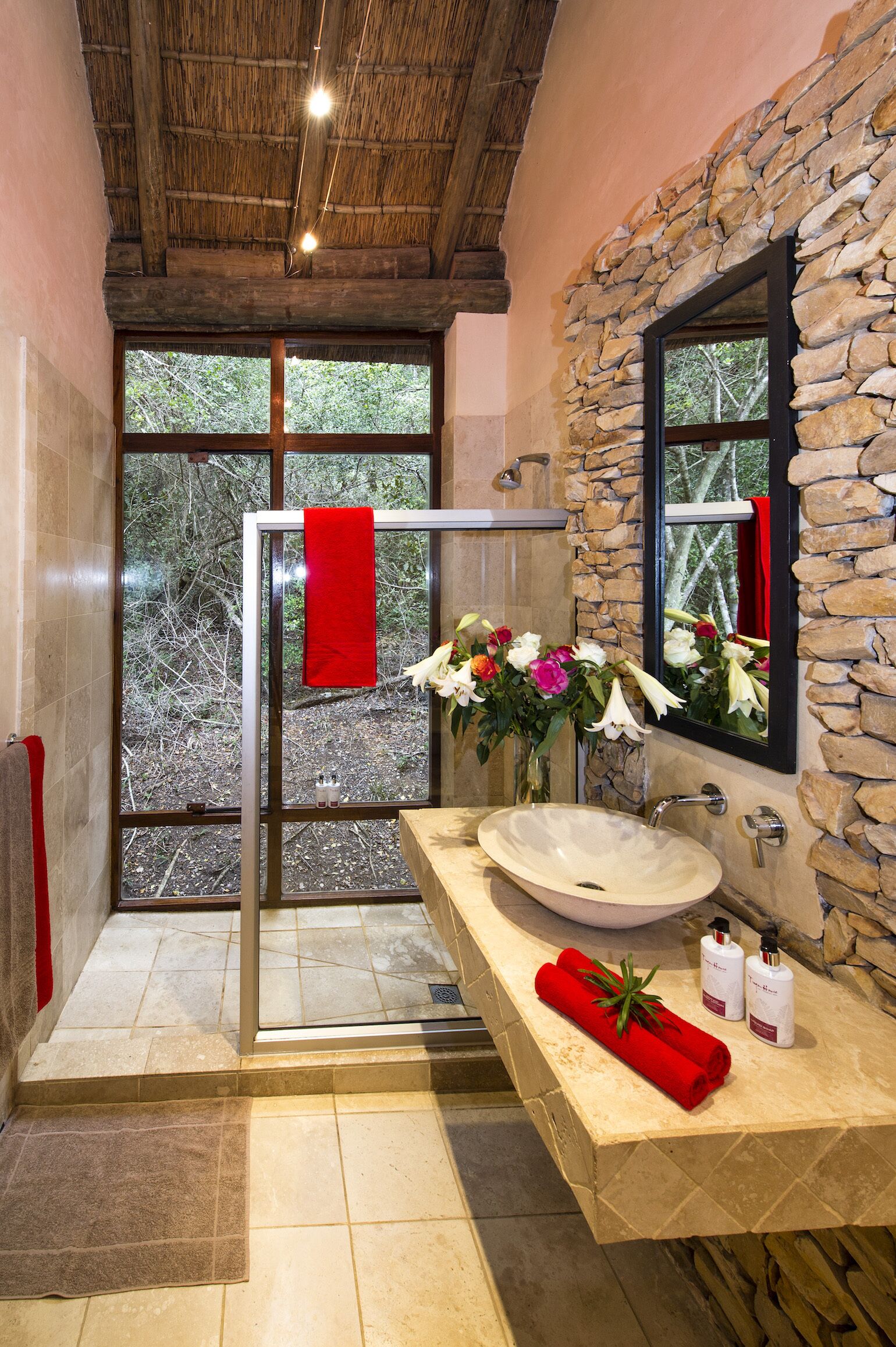 Une salle de bain avec un mur en pierre, une grande fenêtre, une douche en verre et un lavabo beige surmonté d’une serviette rouge.
