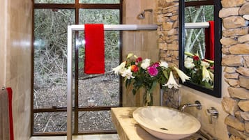 Une salle de bain avec un mur en pierre, une grande fenêtre, une douche en verre et un lavabo beige surmonté d’une serviette rouge.