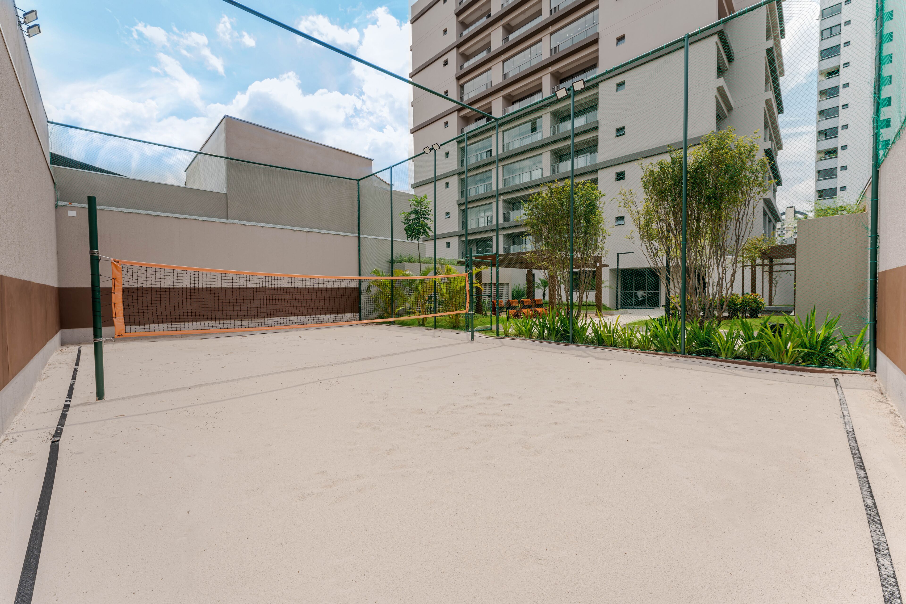 An outdoor tennis court with a net, surrounded by a glass fence, and adjacent to a multi-story building.