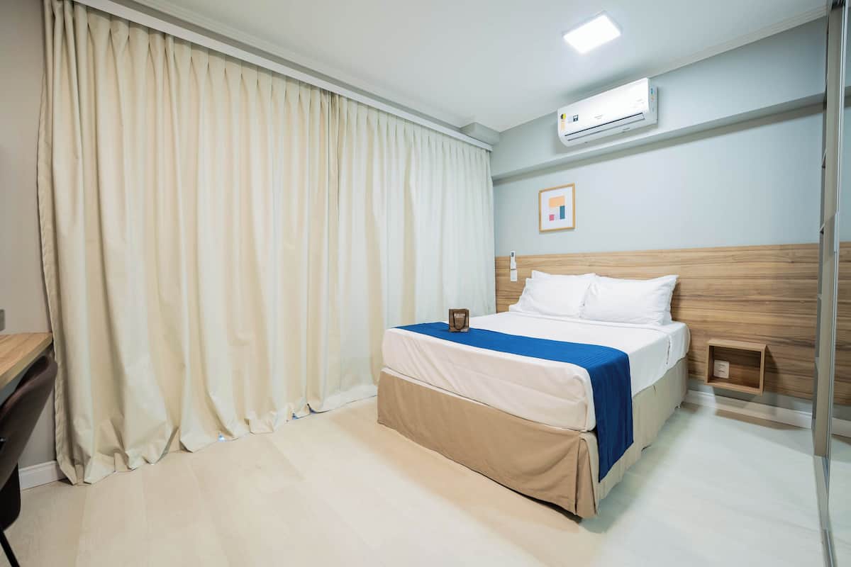 Habitación de hotel con cama, escritorio, silla, mesita de noche, aire acondicionado y ventana con cortinas.